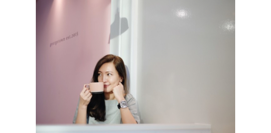 Asian woman standing against a wall ,smelling a cup while grasping her hair lightly with the other hand